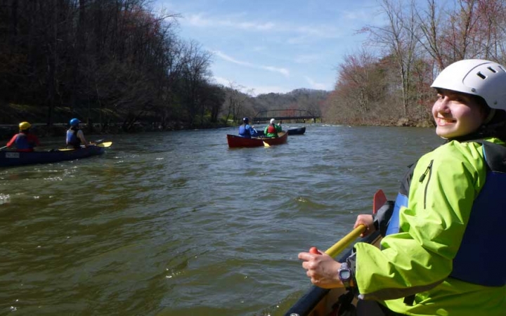 In the foreground, a person sitting at the front of canoe turns around and smiles for a photo. There are other canoes being navigated on the river in the background. 
